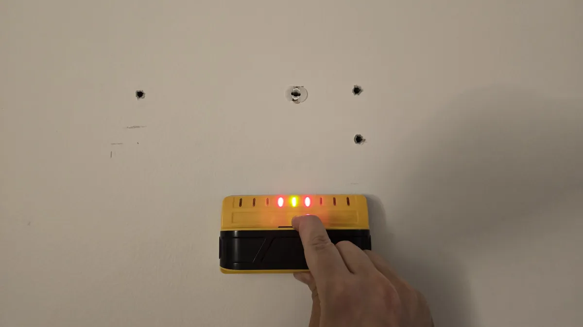 Stud finder lighting up on a drywall wall next to drilled holes