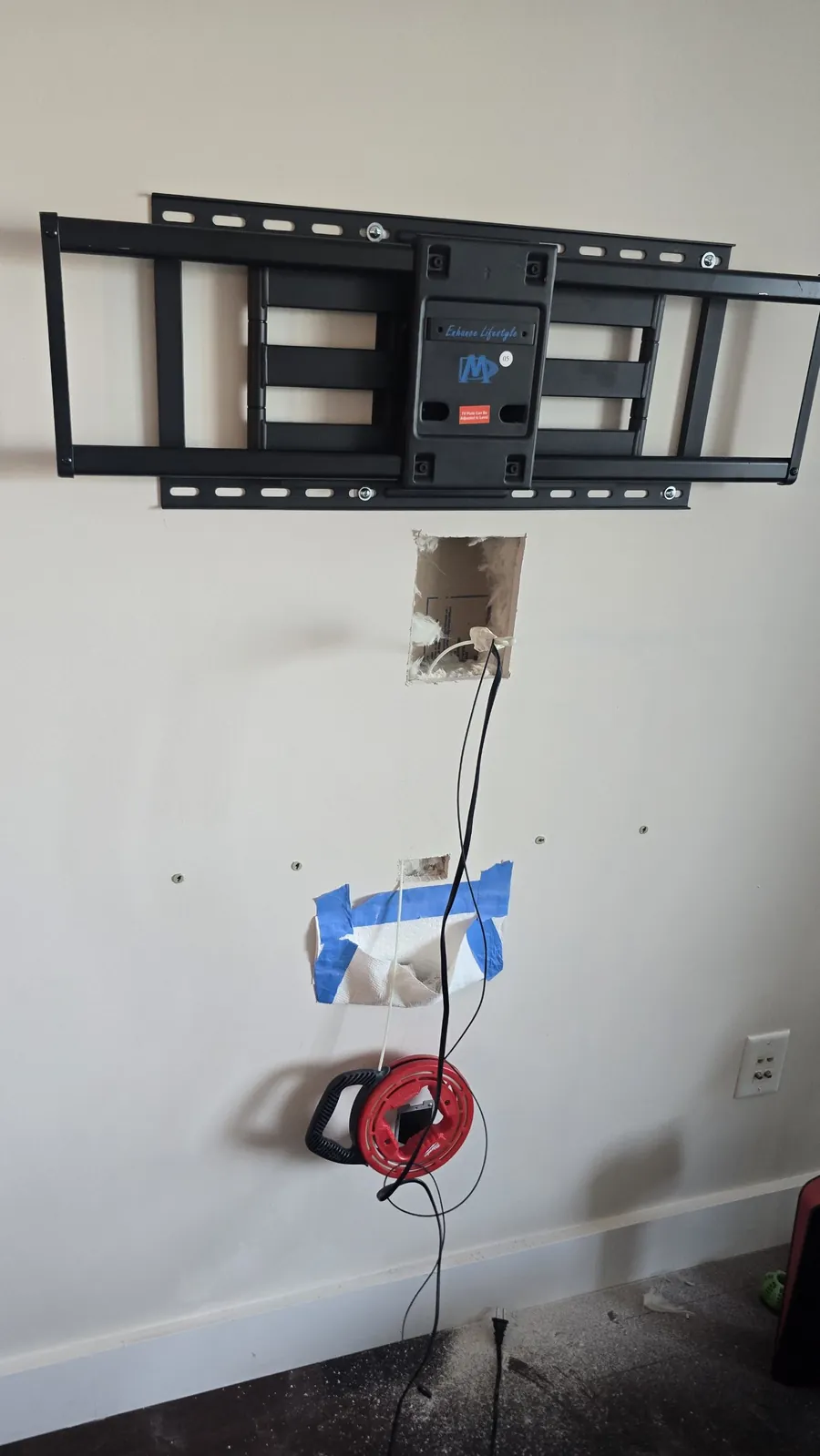 Square openings cut in drywall under a TV mount, with a bag taped below to catch dust and a fish tape reel